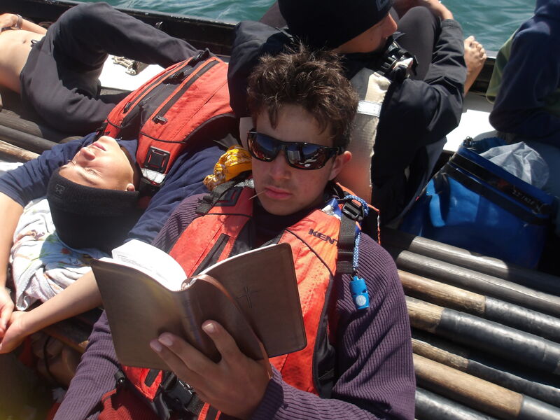 The image shows a group of people on a boat. One person is wearing sunglasses and a life vest while reading a book. Other people are resting or wearing life vests. The setting appears to be outdoors, possibly on a lake or ocean.